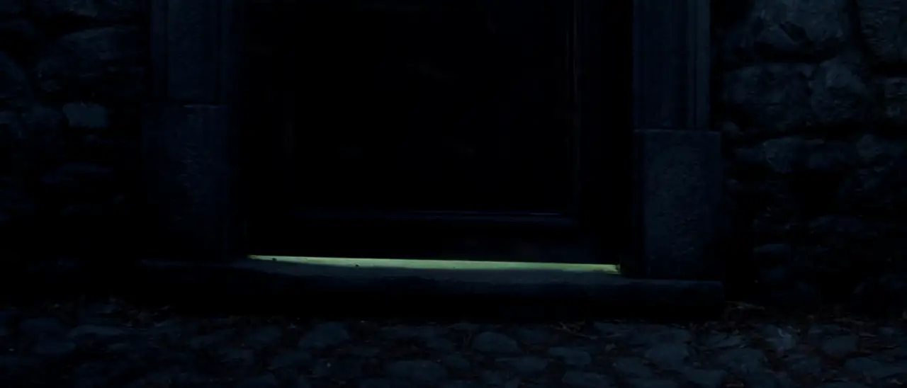 Uma porta de pedra fechada na penumbra, com uma luz fria vazando pela fresta de baixo.